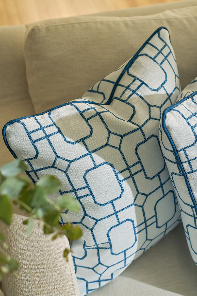 Two Blue Lattice Oversized Pillows featuring coastal blue geometric patterns and piping rest on a light beige sofa, with a green plant partially visible in the lower left corner and warm natural light filling the scene.