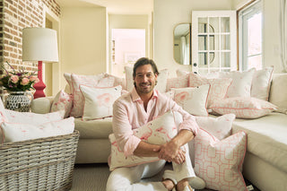 A smiling man in a light pink shirt sits on the floor of a bright living room with a white couch and patterned pink-and-white pillows, including the Flamingo Cluster Extended Lumbar Pillow, adding coastal charm to the space.