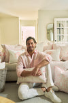 A smiling man in a light pink shirt and white pants sits on the floor, holding The Flamingo Lumbar Pillow. Surrounded by baskets of these vibrant pillows, he brings tropical cheer to the cozy, sunlit living room.