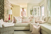 A smiling man in a light pink shirt stands in a sunlit living room with a white sofa decorated with pink and white pillows, including the Flamingo Lumbar Pillow by Sewing Down South, adding tropical cheer to the space.