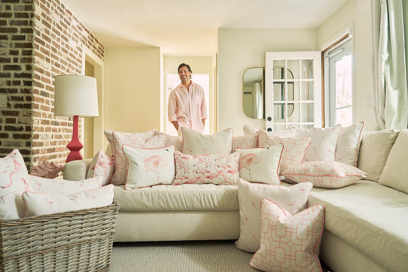 A smiling man in a light pink shirt stands in a sunlit living room with a white sofa decorated with pink and white pillows, including the Flamingo Lumbar Pillow by Sewing Down South, adding tropical cheer to the space.