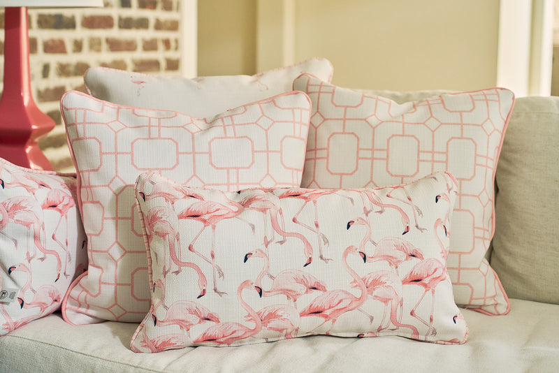 Four decorative pillows add a tropical vibe to a light sofa: two with geometric pink and white designs, and two Flamingo Cluster Extended Lumbar Pillows that pop. A touch of red from a lamp and an exposed brick wall complete the look.