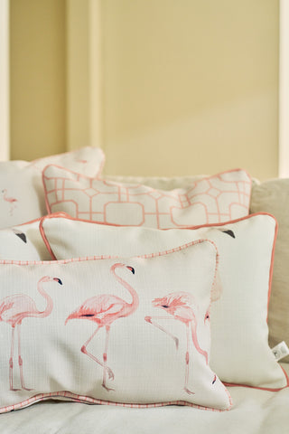 A close-up of decorative pillows on a couch shows the Flamingo Cluster Extended Lumbar Pillow, featuring pink flamingo prints, geometric patterns in soft pastels, peach piping on the edges, and a light coastal look.
