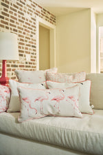 The Flamingo Cluster Extended Lumbar Pillow adds a tropical touch to a light-colored sofa in a cozy living room with exposed brick walls and a table lamp in the background.