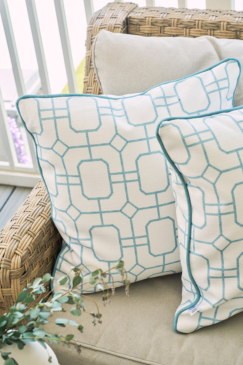 Two Beach Lattice Oversized Pillows featuring a teal geometric design add coastal comfort to a wicker chair with beige cushions. A green leafy plant sits in the foreground, while a railing is visible in the background.