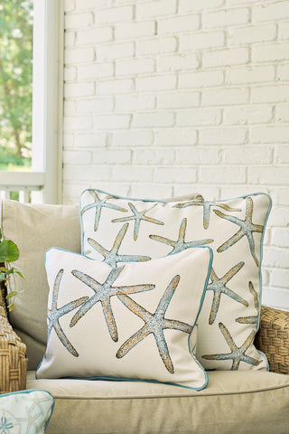 A Starfish Trio with Beach Lattice Lumbar Pillow and another decorative pillow rest on a beige chair with a wicker armrest, featuring blue and tan starfish designs against a white brick wall and greenery background.