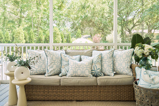 A wicker sofa with beige cushions and blue-and-white pillows, including the Beach Lattice Oversized Pillow for a coastal vibe, sits on a screened porch beside two small tables with greenery vases and a basket holding a white throw.