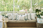 A wicker sofa with beige cushions and blue-and-white pillows, including the Beach Lattice Oversized Pillow for a coastal vibe, sits on a screened porch beside two small tables with greenery vases and a basket holding a white throw.