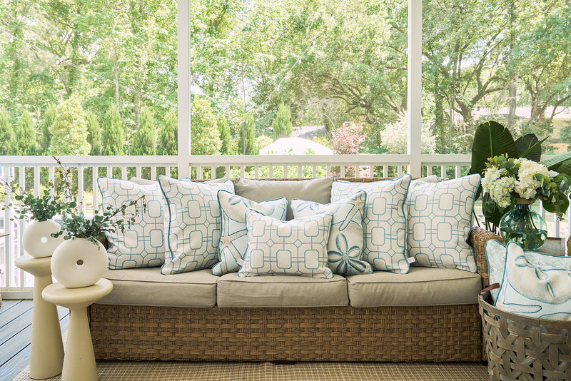 A wicker sofa with beige cushions and blue-and-white pillows, including the Beach Lattice Oversized Pillow for a coastal vibe, sits on a screened porch beside two small tables with greenery vases and a basket holding a white throw.