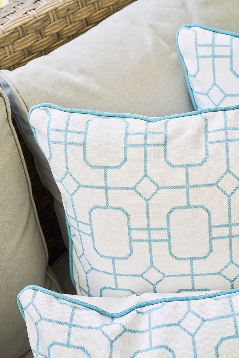 The Beach Lattice Oversized Pillow, featuring a light blue geometric print and blue edging, brings a coastal vibe to a light gray cushioned seat with a woven wicker armrest in view.