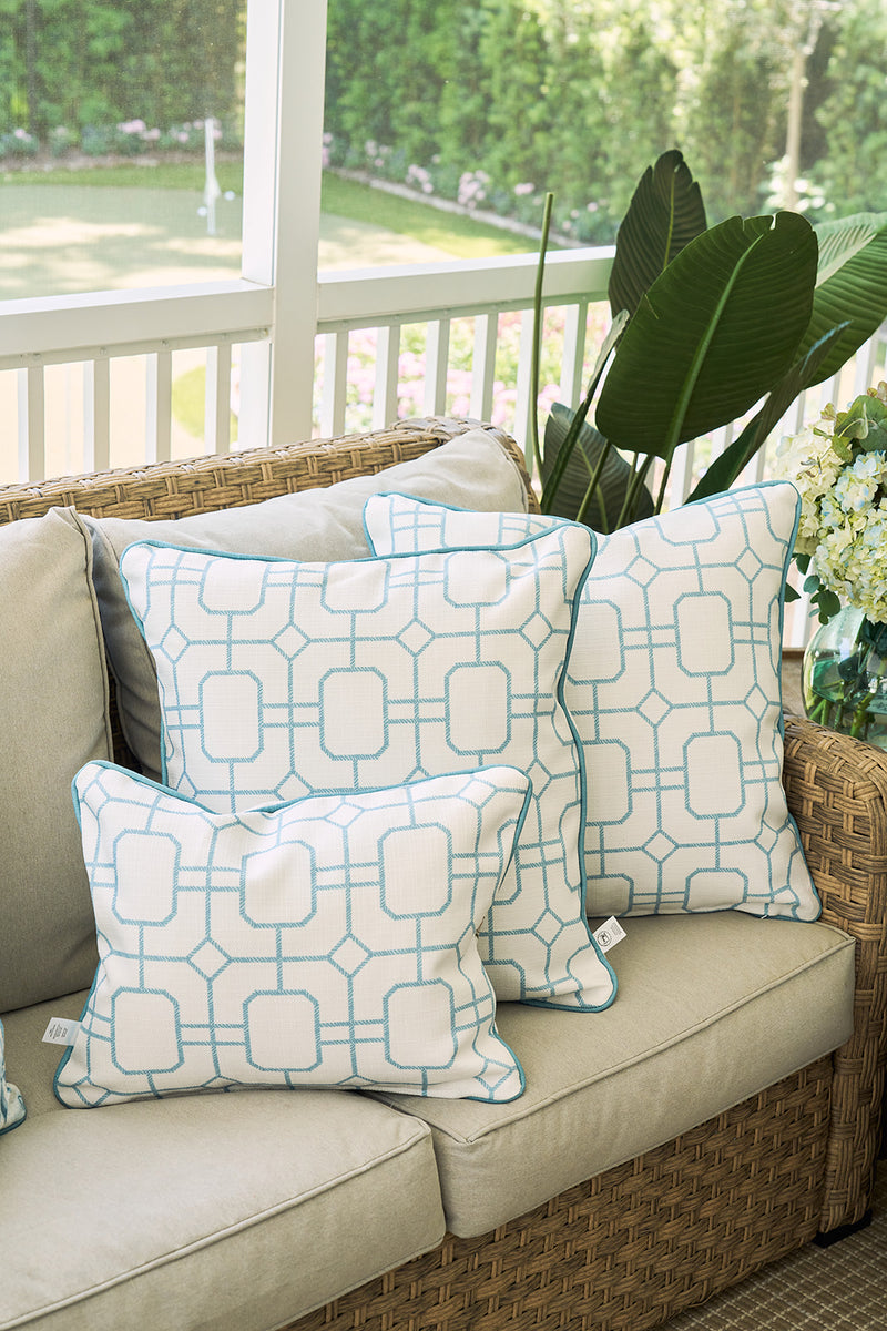 The Beach Lattice Oversized Pillow, with its blue geometric pattern, brings a subtle coastal touch to your space when styled on a beige outdoor sofa amid green plants and a white railing.