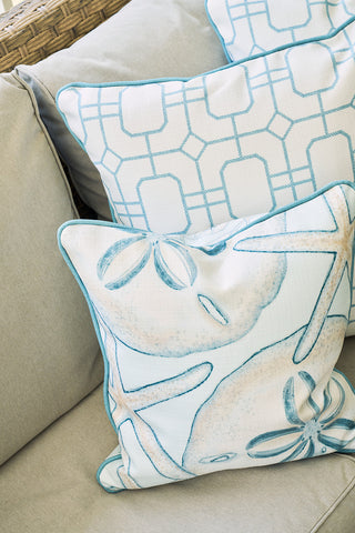 Three decorative pillows sit on a beige sofa for coastal flair. Two have light blue geometrics, while The Beaches Pillow in front features sand dollar and starfish designs in soft blue tones.