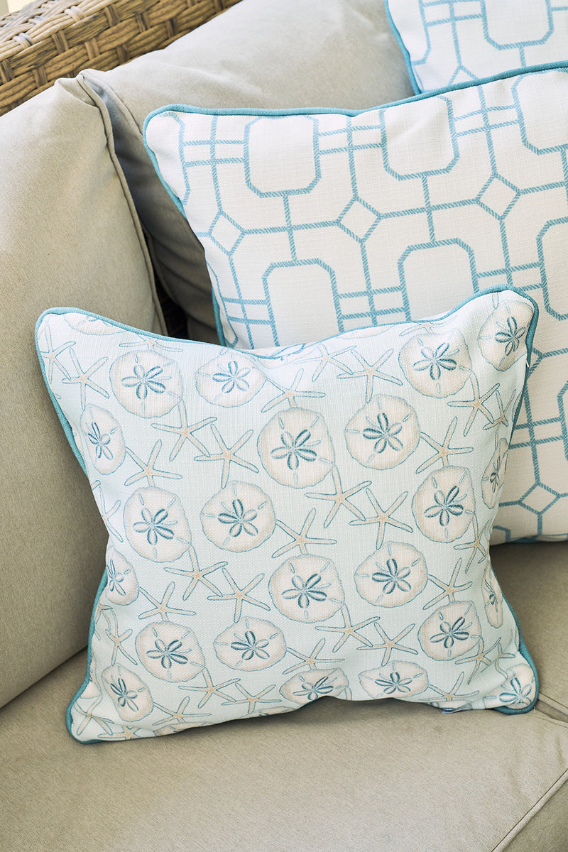 The Beaches Pillow features a light blue and beige sand dollar design, displayed on a beige couch beside a blue and white geometric pillow—ideal for bringing coastal decor to any living space.
