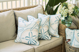 Beige outdoor sofa with three The Beaches Pillows, featuring starfish and sand dollar designs, evoking seaside nostalgia next to a wicker table topped with white flowers and green leaves.