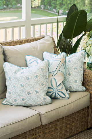 Three The Beaches Pillow in light blue, featuring sea-inspired patterns like starfish and sand dollars, adorn a beige wicker sofa, creating relaxed coastal decor on a porch overlooking the green yard.