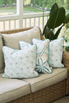 Three The Beaches Pillow in light blue, featuring sea-inspired patterns like starfish and sand dollars, adorn a beige wicker sofa, creating relaxed coastal decor on a porch overlooking the green yard.