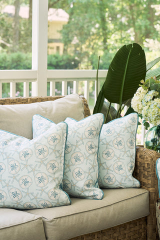 A wicker outdoor sofa with light beige cushions and three The Beaches Pillow, featuring a blue starfish pattern. Green leaves and a vase of white flowers complete the coastal look, with a porch railing and trees behind.