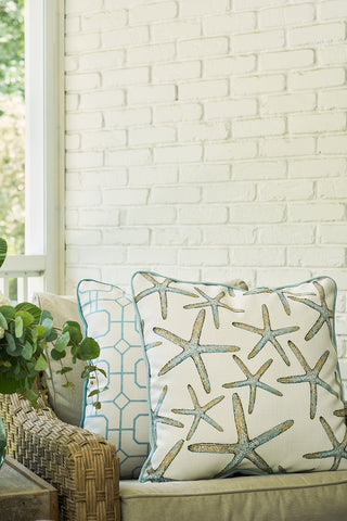 Two decorative pillows on a wicker chair bring coastal chic to your space: one is the Starfish Oversized Pillow, while the other has a blue geometric design. A green potted plant and white brick wall complete the look.