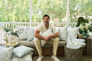 A smiling man sits on a cushioned outdoor bench, holding a Starfish Trio with Beach Lattice Lumbar Pillow. Matching pillows and lush greenery through large windows create a cozy, bright, ocean-inspired atmosphere.