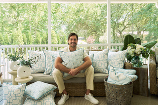 A man smiles on a sofa accented with The Beaches Pillow, featuring white and light blue coastal patterns, on a sunlit porch with lush greenery visible through large windows.