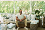 A man smiles on a sofa accented with The Beaches Pillow, featuring white and light blue coastal patterns, on a sunlit porch with lush greenery visible through large windows.