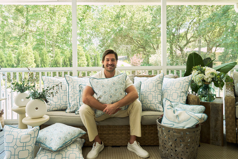 A man smiles on a sofa accented with The Beaches Pillow, featuring white and light blue coastal patterns, on a sunlit porch with lush greenery visible through large windows.