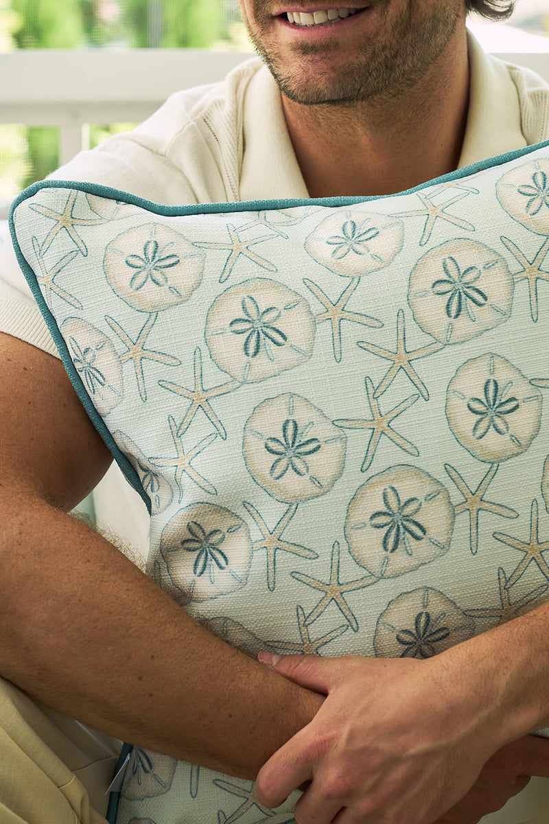A man hugs The Beaches Pillow, featuring sand dollar and starfish patterns, while sitting indoors against a blurred green backdrop, evoking seaside nostalgia.
