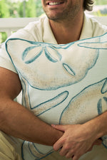 A man in a white shirt smiles while hugging The Beaches Pillow, featuring large blue-outlined sand dollar designs. Green foliage appears in the blurred background, highlighting the pillows coastal decor vibe.