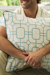 A man smiles, sitting with his arms wrapped around the Beach Lattice Oversized Pillow, which features white fabric with a bold blue geometric lattice pattern and blue piping along the edges.