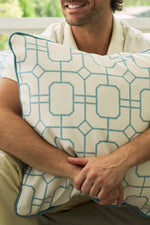A man smiles, sitting with his arms wrapped around the Beach Lattice Oversized Pillow, which features white fabric with a bold blue geometric lattice pattern and blue piping along the edges.