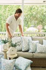 A man in a white shirt arranges Beach Lattice Oversized Pillows on an outdoor wicker sofa, surrounded by lush greenery. A nearby table displays sea-glass-hued vases filled with greenery.