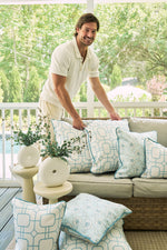 A smiling man arranges the Beach Lattice Oversized Pillow and other blue-and-white pillows on a wicker sofa on a sunny porch, surrounded by greenery, vases, coastal decor, and with a swimming pool visible in the background.