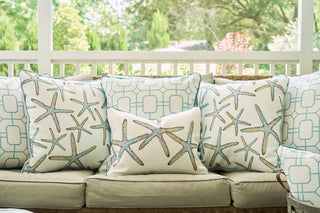 Coastal chic outdoor sofa features the Starfish Oversized Pillow and blue geometric Sewing Down South pattern pillows, set on a porch with lush green views and a white railing.