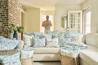 A man stands in a bright coastal-themed living room holding a wreath. The space features a white sofa and blue-and-white pillows, including The Sandpiper Pillow, with extra pillows in baskets, plus a brick wall and flowers.