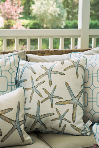 Decorative pillows on an outdoor sofa feature the Starfish Oversized Pillow by Sewing Down South™ in blue and tan. Geometric patterned pillows and lush greenery create a coastal chic vibe in the background.