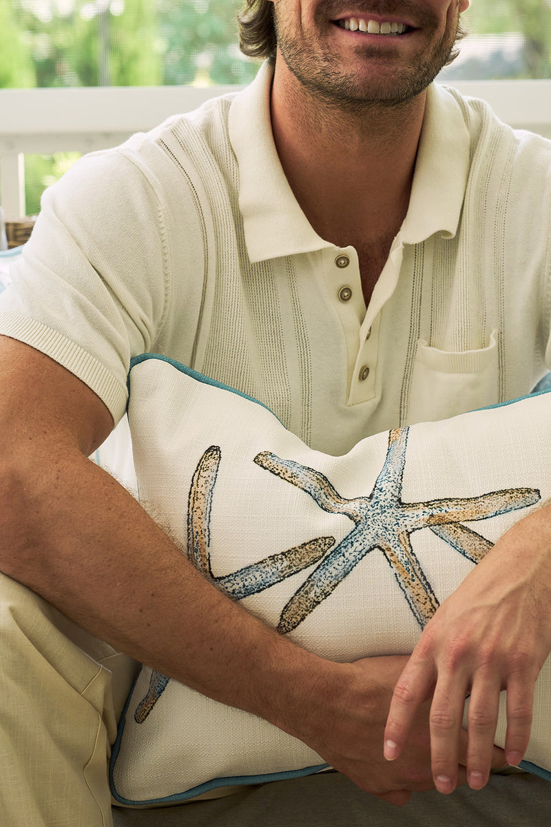 A man in a cream-colored shirt sits on a couch, smiling and holding the Starfish Trio with Beach Lattice Lumbar Pillow. Softly blurred greenery in the background adds to the scenes coastal style and ocean-inspired vibe.