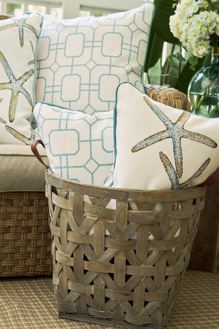 A woven basket holds decorative pillows, including a Starfish Trio with Beach Lattice Lumbar Pillow and a blue geometric one. More coastal pillows accent a nearby wicker couch, while a vase of white flowers brightens the background.