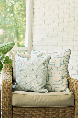 A cozy wicker chair with light tan cushions features two of The Beaches Pillow in blue and white patterns, set against a white brick wall and greenery for a relaxed, coastal vibe.