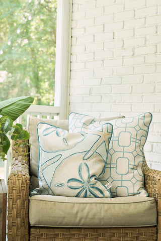 A wicker chair with beige cushions sits on a porch. Two decorative pillows, including The Beaches Pillow with sand dollar designs, evoke seaside nostalgia. Green plants and a white brick wall are in the background.