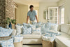 A man stands behind a cream sofa adorned with blue and white pillows, including the Blue Lattice Oversized Pillow. Exposed brick and bright windows enhance the cozy, sunlit room’s coastal blue style.
