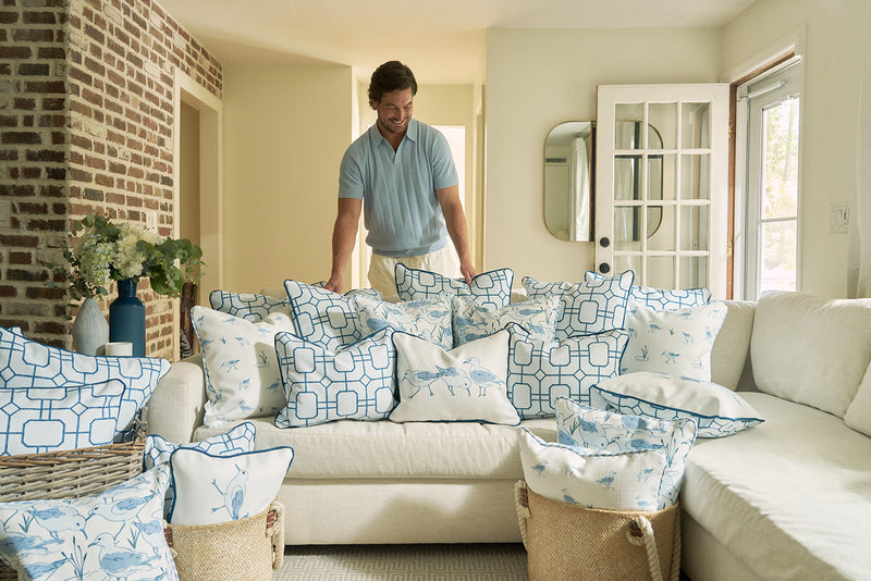 A man stands behind a cream sofa adorned with blue and white pillows, including the Blue Lattice Oversized Pillow. Exposed brick and bright windows enhance the cozy, sunlit room’s coastal blue style.