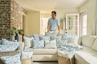 A man stands behind a cream sofa filled with white and blue pillows, including the Sandpiper Trio with Blue Lattice Lumbar Pillow, in a bright living room with brick walls, wicker baskets, and floral decor for a beachy vibe.