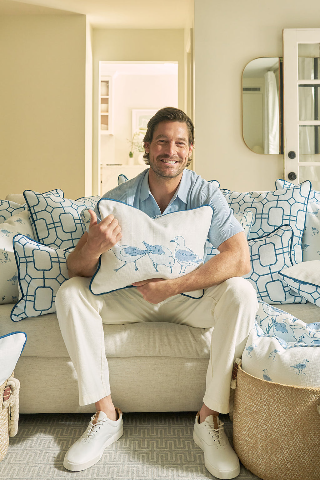 A man sits on a cream sofa, smiling and giving a thumbs up while holding a Sandpiper Trio with Blue Lattice Lumbar Pillow. The sofas blue and white patterned pillows enhance the cozy, beachy vibe of the neutral room.