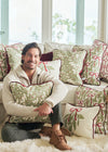 A man sits on a white couch hugging a Jolly Holly Pillow, surrounded by decorative holiday pillows with red and green patterns, in a bright room with Christmas decorations.