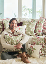 A man sits on a white couch hugging a Jolly Holly Pillow, surrounded by decorative holiday pillows with red and green patterns, in a bright room with Christmas decorations.