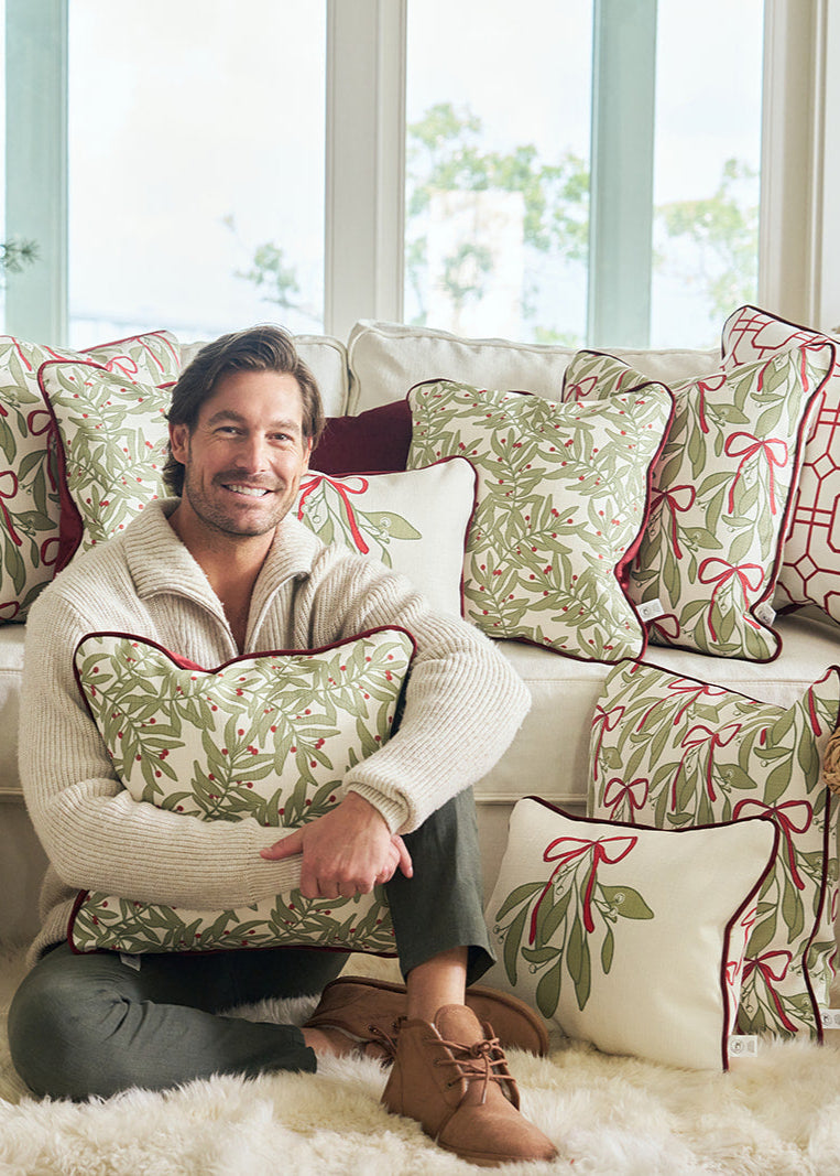 A man sits on a white couch hugging a Jolly Holly Pillow, surrounded by decorative holiday pillows with red and green patterns, in a bright room with Christmas decorations.