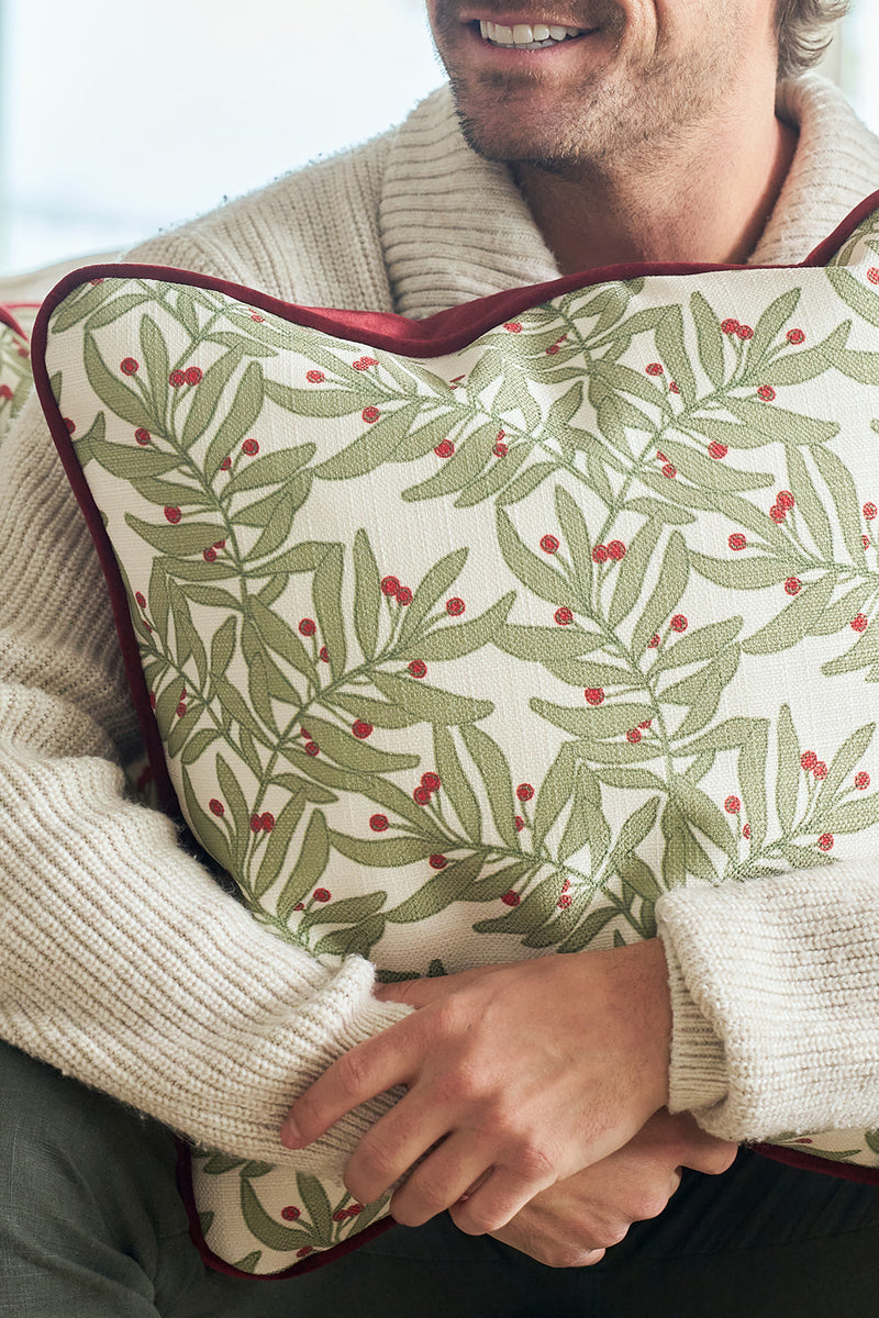 A smiling man in a cream knit sweater hugs the Jolly Holly Pillow, which features a green leafy and red berry pattern with dark red piping.