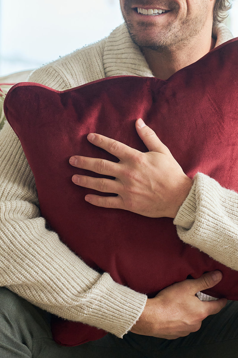 A man in a cream sweater smiles and hugs the Jolly Holly Pillow, a large, red velvet holiday cushion, with only the lower half of his face shown.