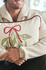 A man in a beige sweater smiles while hugging the Bows & Mistletoe Lumbar Pillow, which features charming mistletoe and red ribbon accents.