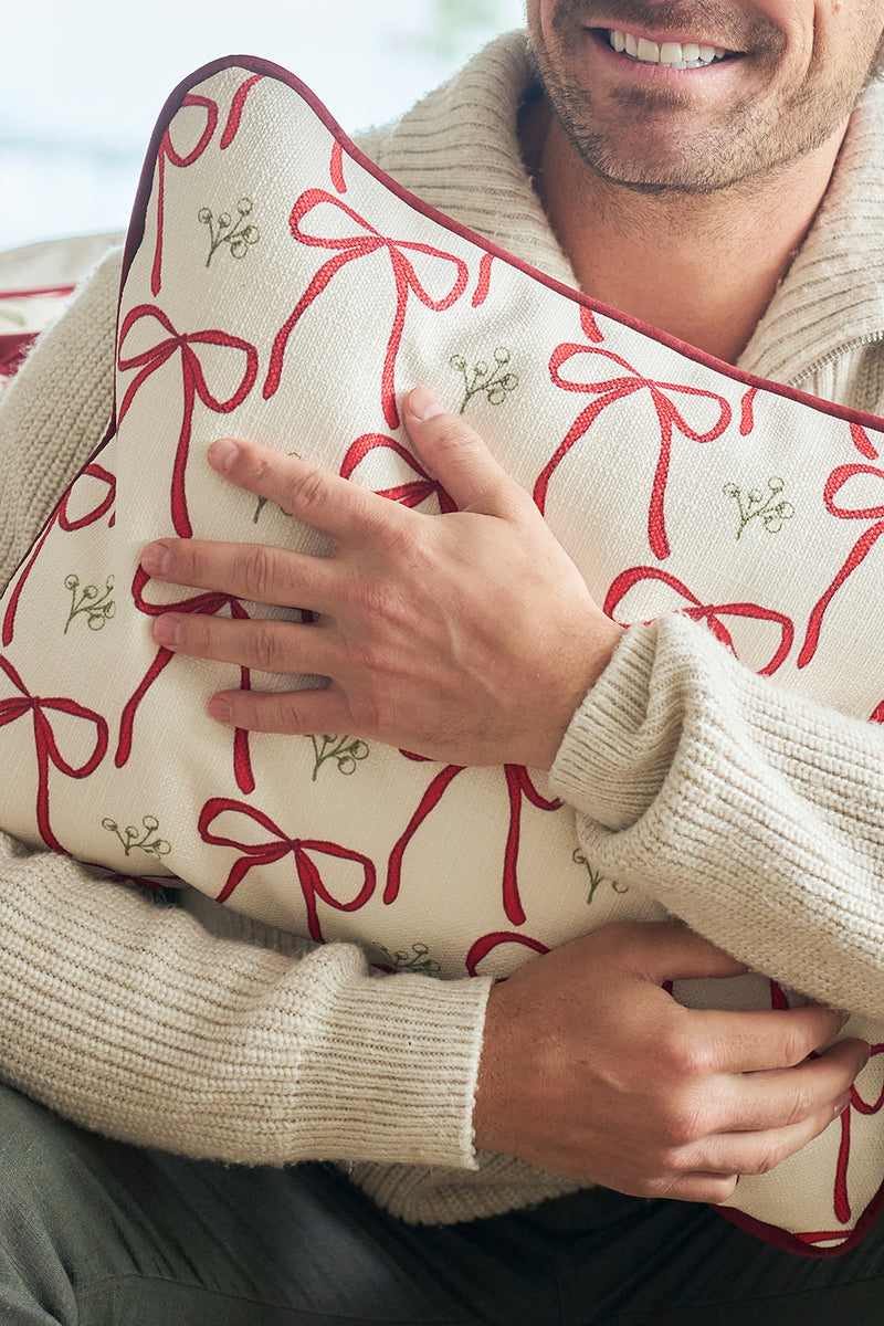 A person in a beige sweater holds the Bows & Mistletoe Lumbar Pillow, featuring festive red ribbon designs and green mistletoe accents, while smiling gently.
